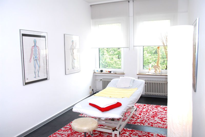Behandlungsraum mit Liege, Hocker und Anatomietafeln an der Wand